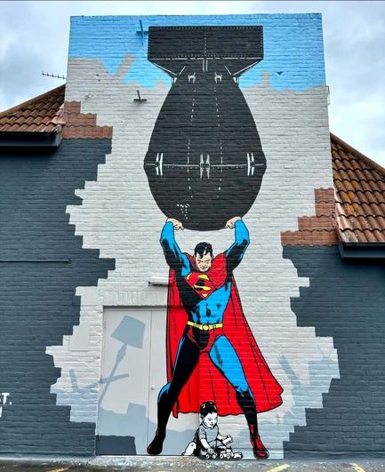 Streetartwall. A large mural of Superman was spray-painted on a one-story exterior wall between the roofs of a rooftop parking lot. Superman stands with his amen facing upwards, holding up a giant bomb. He is looking down as a small baby sits between his legs on the ground playing with his toy cars. (Superman is depicted in blue and red, the child in gray/black/white).
Info: The artist says:  
"There's room at the top they are telling you still, but first you must learn how to smile as you kill" - John Lennon.
