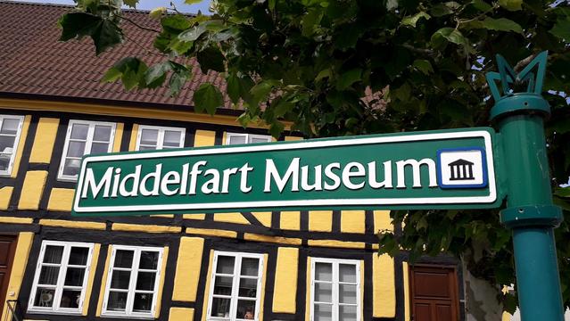 Beyond, framed by leafy tree branches, a section of a medieval half-timbered brick building in the lovely town of Middelfart, Denmark. It is painted a lovely buttercup yellow, crisscrossed by thick black planks of timber. There are two rows of multi-pane windows with white frames. To our right, a tall metal green pole. From it, a fingerpost sign points in across the whole image, filling the foreground in front of our eyes. It is elegant. Deep green with a white border. To the right edge: that little international sign for 'museum'. The one I assume was designed with a Western European museum filled with colonial loot in mind, because it's a line drawing of a Greek temple (with no marbles). The sign directs us to Middelfart's excellent local history museum. It's the kind of sign people enjoy reading with English-speaking eyes. Middelfart Museum. The Museum of Middle Farts. If it were an art museum, it would be even more fun for our inner teenager to say in English. The Danish for art is: Kunst. Middelfart Kunstmuseum.