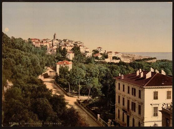 The image depicts a coastal town, possibly in the Italian Riviera. In the foreground, there are houses with visible architectural features such as balconies and windows. A street runs alongside these buildings, suggesting a residential area close to the sea. There is also vegetation like trees and shrubs, contributing to the picturesque nature of the scene. The background shows a glimpse of the coastline and the water, with boats visible on the sea, indicating that this town may be involved in maritime activities such as fishing or tourism. The sky is clear, suggesting a bright day, which adds to the overall serene and idyllic atmosphere of the image.