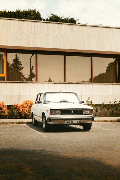 The image shows an older model car parked in what appears to be a public parking space. The vehicle has a distinctive boxy shape, which is characteristic of some cars from the 1970s or 1980s. There's no visible text on the car or nearby buildings to provide context about its make or model. The setting looks like an urban environment with concrete surfaces and what might be
part of a sidewalk or walkway.