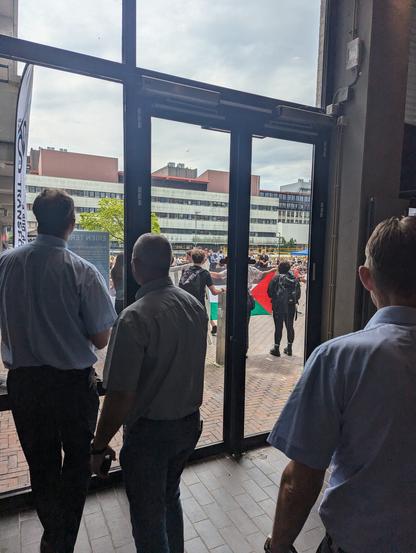 Picture from the inside of a campus building looking through large windows and seeing Pro-Palestine protest outside, a flag. There's security men in front of the doors. No way out