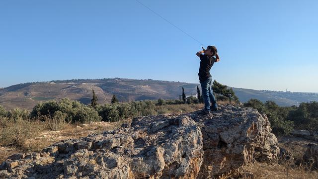 Child standing on a rock maneuvering a kite, with a blue sky and beautiful hills in the background