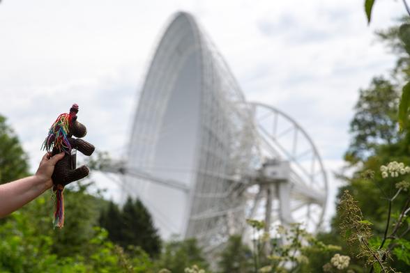 Das Bild zeigt Max Horn, das bunte Strickhorn, in der linken unteren Ecke des Bildes, der in die Richtung eines beeindruckenden Radioteleskops schaut. Das Radioteleskop Effelsberg erhebt sich majestätisch in die Höhe, sein riesiger Parabolspiegel dominiert die Szene. Der Himmel ist leicht bewölkt, was einen Kontrast zur weißen Struktur des Teleskops bildet. Umgeben von Bäumen und Grünflächen, scheint das Teleskop aus der Natur herauszuwachsen. Max Horn wird von einer Hand gehalten und wirkt fasziniert von der technischen Konstruktion vor ihm. [ChatGPT4o]

In diesem Bild liegt die Schärfe auf Max, die große Schüssel verschwimmt unscharf im Hintergrund.