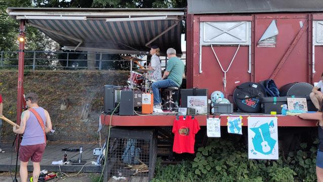 Der Waggon in Offenbach. Ein alter, roter Güterwaggon in dem Konzerte stattfinden. Low Budgie spielen draußen. Bass und Drums oben auf der Plattform, der Girarrist unten auf dem Gleis. Am Waggon hängt außerdem Merch (Poster, Shirt, Platte, CD)