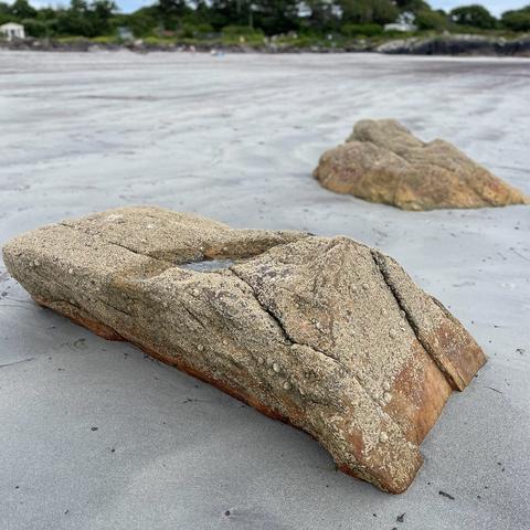 A close-up of large weathered rocks on a sandy beach with distant greenery in the background.