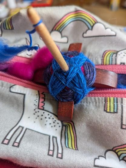 A cross arm drop spindle with blue and pink fiber in a unicorn themed bag under it.