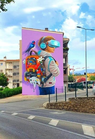 Streetartwall. A beautiful mural of a little boy with a backpack was sprayed/painted on the exterior wall of a three-story modern apartment building. The background is in pink. The brown-haired boy is wearing a light-colored hoodie sweater, jeans, headphones and 3D glasses. He has a red rucksack on his back filled with all kinds of electronics and toys. Inside is a Nintendo controller, a Batman pendant and a pencil. There are also two white flowers, a spray can, lots of buttons, a small speaker box and much more. A blue and white bird sits enthroned on the handle of the rucksack. A beautifully designed mural with lots of charm.