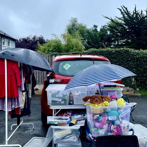 A yard sale setup in front of a red car. Items for sale include clothing on a rack and various toys in plastic bins. The setup is covered by umbrellas to protect it from the rain.