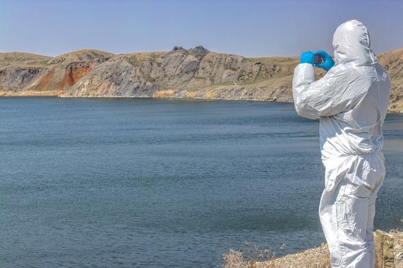 A man in a white #HAZMAT suit stands before a large blue lake and takes a photo