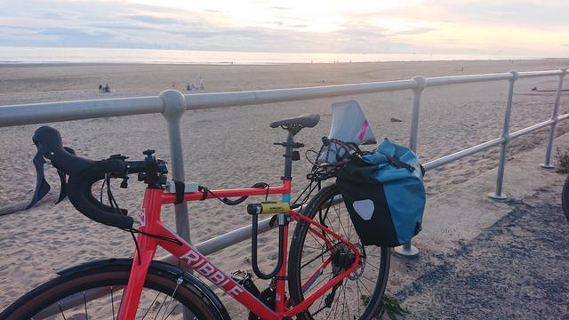 A gravel bike with a pannier and a megaphone mounted on the back leans against railings at the edge of the beach. People are dotted along the sand and in the water. The sun is setting and shining through light cloud
