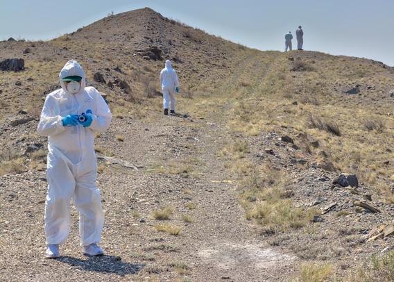 A man in white HAZMAT suit stands in the rough terrain surrounding the Atomic Lake. Some other figures in the back ground climb a hill.