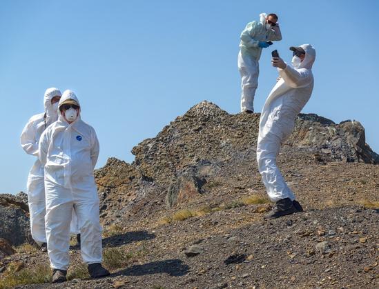 A group of people in HAZMAT suits stand on a a heap of earth. One holds a mobile phone ready to take a selfie.