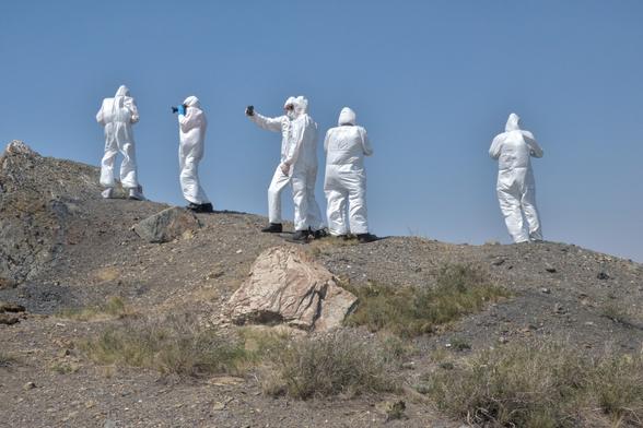 A group of people in HAZMAT suits stand on a a heap of earth. One holds a mobile phone ready to take a selfie.