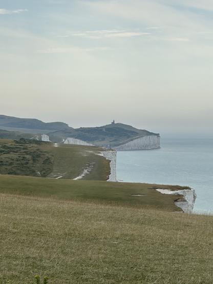 The view from the western end of the Seven Sisters over to Beachy Head in the East, with the sea on the right and green slopes on the left. The sky is a hazy blue.