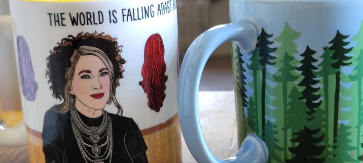 Two mugs, one is Moira Schitt before her wall of wigs, the other a conifer forest with blue background. These are the dides that face away from a right-handed user