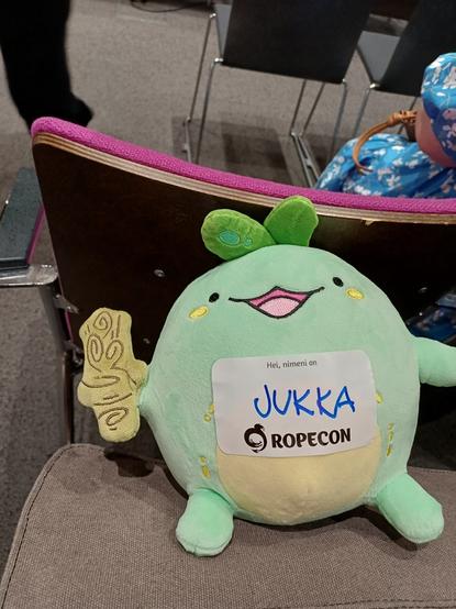 Iloisesti hymyilevä miekkaa pitelevä vihreä Quest Sprout -pehmolelu, jonka masuun on kiinnitetty "Hei, nimeni on Jukka" -tarra Ropecon-logolla.