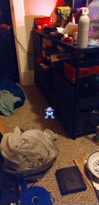 A glitched Pokémon Go AR screenshot with a very tiny Popplio next to a small drawer, in a cluttered bedroom.