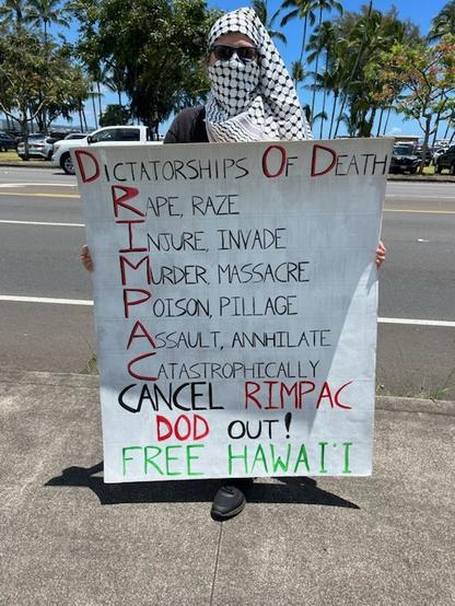Person wearing a kufiya holding a sign that says:
D O D dictatorships of death.
The letters in RIMPAC are used to begin the words: Rape, Raze, Injure, Invade, Murder, Massacre, Poison, Pillage, Assault, Annihilate, Catastrophically Cancel RIMPAC Department Of Defense OUT! Free Hawai'i!