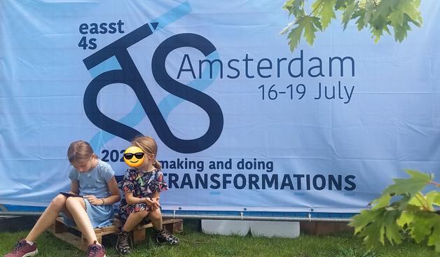 Two kids sitting in the shade of a banner of the EASST4S conference. Banner says "making and doing transformations". One kid is reading, the other looks aside.