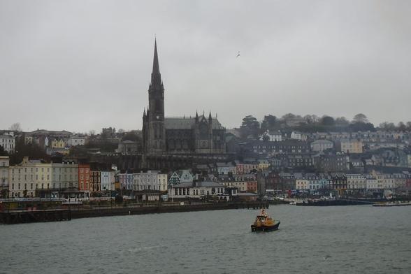 A view of Cobh from the sea. The Cork Pilot boat is in the foreground. St. Colman's Cathedral stands tall against a grey sky. Colourful houses run along the harbour.