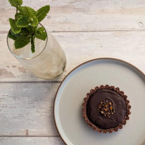 A square photo of an individual chocolate tart on a plate, sat next to a mint julep cocktail in a highball glass. The tart has a dark chocolate biscuit crumb case, and is filled with a dark chocolate ganache; on top are some wee brown crystals of boiled sweets. The mint julep is a light-brown liquid over crushed ice, with mint spring garnish. They sit on a white wooden background.