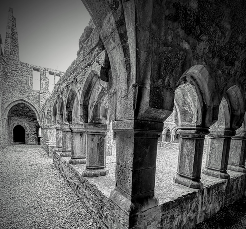 A black and white photo of a corner in a ruined abbey’s colonnade, with an open doorway at the end of one of the wings.