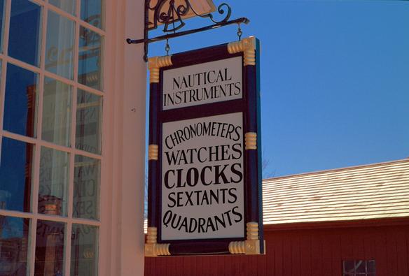 Color photo of a hand crafted wooden sign with black trim, carved gold painted trim, and old style caps black lettering on a white background. The top section of the sign reads: Nautical Instruments. The bottom section reads: Chronometers, Watches, Clocks, Sextants, Quadrants. The words Chronometers and Quadrant arc above and below the other words in the bottom section. The sign hangs from a wooden paned display window of a reconstructed instrument shop at Mystic Seaport Museum, Mystic CT.