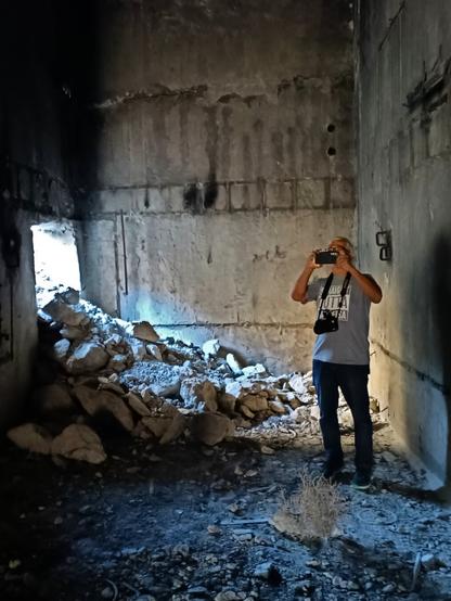 A man takes a photo from inside a nuclear bunker. Rubble has fallen in through an opening in the wall behind him.