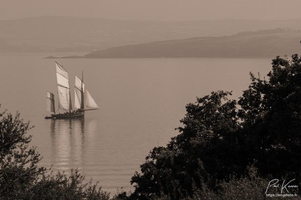 Photo monochrome, sépia présentant un vieux gréement, la bisquine Cancalaise, naviguant par petit temps sur une mer d'huile où se reflètent la voilure et les trois mâts du bateau.
La photo est prise depuis le rivage, un peu en hauteur, par dessus la cime des arbres que l'on voit au bas de la composition alors qu'au haut de la photo, en arrière-plan, on distingue dans la brume, une côte également élevée.