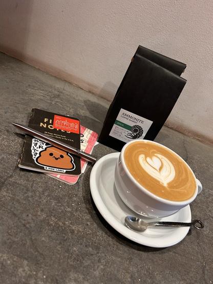 latte with heart-shaped florets in a white cup (with a saucer and small spoon), a couple of Field Notes notebooks, a pen, and a bag of coffee beans