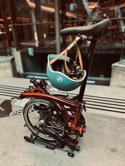 Formato vertical de mi Brompton lacada naranja plegada, pero con el sillín extendido. De él cuelga el casco azul. Y de fondo se ve la cristalera de la piscina de submarinismo. El suelo es como de cementaco y todo el centro comercial es industrial.