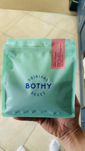 A hand holding a bag of Bothy Roast coffee beans. The label indicates the coffee's origin as Nariño, Colombia, and describes its flavors as honey caramel and chocolate with citrus fruits. The bag is light blue with blue and pink printing.
