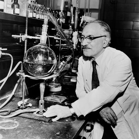 A portrait of Selman A Waksman in a laboratory in front of laboratory equipment. He is wearing a laboratory coat, a light shirt and tie, glasses and a moustache.