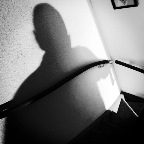 Black and white picture showing the shadow of a man on the wall of a staircase. The shadow is cast on the wall by a flash. The railing  is clearly visible, even in the shadow. On the right a small part of a painting is visible. The atmosphere of the photo is a bit eerie.