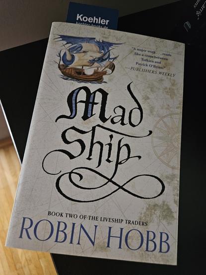 Das Buch auf einem schwarzen Beistelltisch. Das Cover zeigt die Miniatur einer mittelalterlichen Kogge (o.ä.), um deren Masten sich ein blauer Drachen windet. Dieser erinnert irgendwie an die Seeschlangen aus Band 1, ist aber mit Schwingen ausgestattet. Daneben ein Endorsement der Publishers Weekly, die das Werk als Kreuzung zw. Patrick O'Brian (Autor der Aubrey-Maturin-Romane) und Tolkien (Der Herr der Ringe) einordnet.