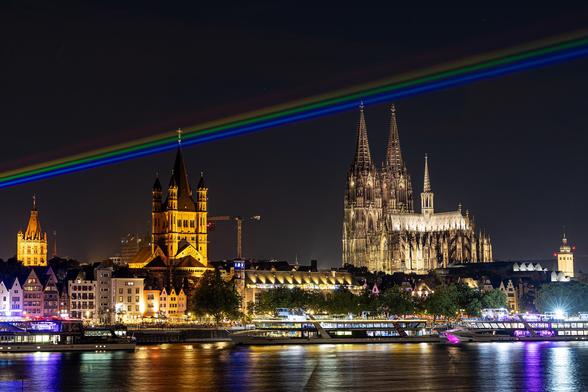 nächtliches Kölner Rheinpanorama  mit Rathaus, Groß-St-Martin, Kölner Dom im Hintergrund, den Altstadthäusern in der Mitte und dem Fluß im Vordergrund.Durch das Turmkreuz von St. Martin und die linke Domspitze verläuft ein in dien Nachthimmel laserprojizierter Regenbogen in Linienform