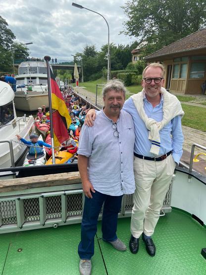 Ralf Dimmek und Eric Oehlmann an Bord der CANOW in der Schleuse Diemitz. Im Hintergrund sind zahlreiche Paddler und Sportboote zu sehen.