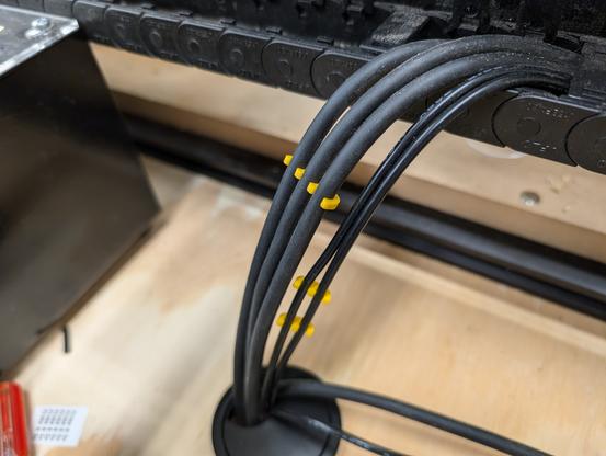 Photo of three large cables, and two small cables exiting a cable chain and entering a cable passthrough. The large cables and small cables are (separately) connected by plastic cable combs in a contrasting color