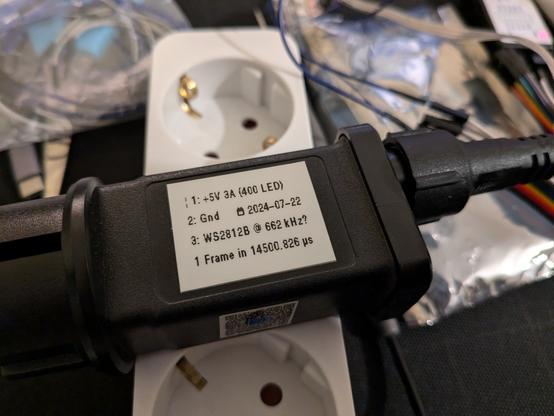 Black integrated power supply and smart LED driver lying on a messy desk on top of a Schuko power strip. It has a white printed sticky label that says:

¦ 1: +5V 3A (400 LED)
2: Gnd    📅 2024-07-22
3: WS2812B @ 662 kHz?
1 Frame in 14500.826 µs
