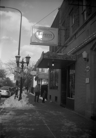 Black-and-white photo of city street scene, signage of a bar, a man and child on the sidewalk with snow.