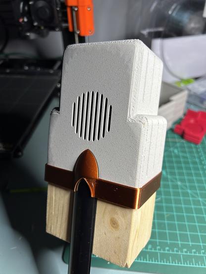 An assembly.  At the bottom, a short unpainted pine board on end, with a black aluminum channel for LEDs embedded in the front side.  At the top, a printed cap with slots for a speaker.  The cap has a rough texture and a sort of art deco tower shape.  Holding them together is a horizontal band of shiny copper printed PLA with a bullet shape.