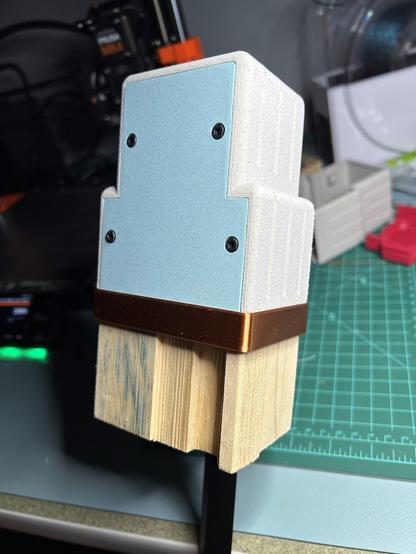 The same assembly, back side. On this side, a channel is seen cut in the board. And the cap has a cover plate in a contrasting color of marble PLA, slate grey, with four hex cap machine screws holding it on.
There is some kind of blue smudge on the board. Various bits of 3D printing tools and detritus are out of focus in the background.