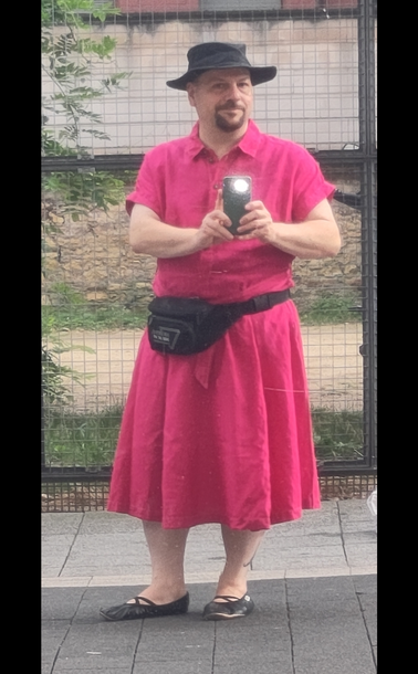 Selbstporträt in spiegelnder Scheibe. Mann mittleren Alters in einem pinkfarbenen Leinenkleid. Das Kleid ist oben geschnitten wie ein Kurzarm-Hemd mit Kragen und Knopfleiste. Der Rockteil ist wadenlang und weit ausgestellt. Dazu trage ich einen schwarzen Hut, eine schwarze Bauchtasche und schwarze Ballerinas.