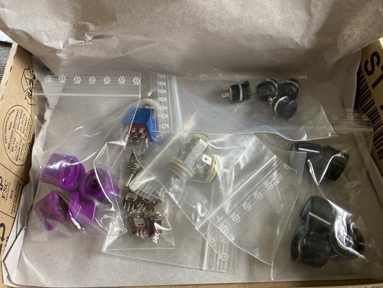 Shipping box with some components for guitar effects pedals. A 3PDT footswitch, some jacks for power and audio, some small switches and a lot of potentiometer knobs.