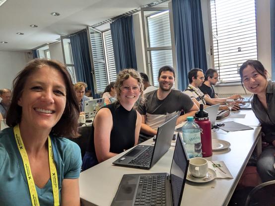 Groups of 2-5 individuals of diverse genders working sitting at long tables working on laptops.