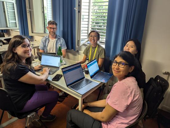Groups of 2-5 individuals of diverse genders working sitting at long tables working on laptops.