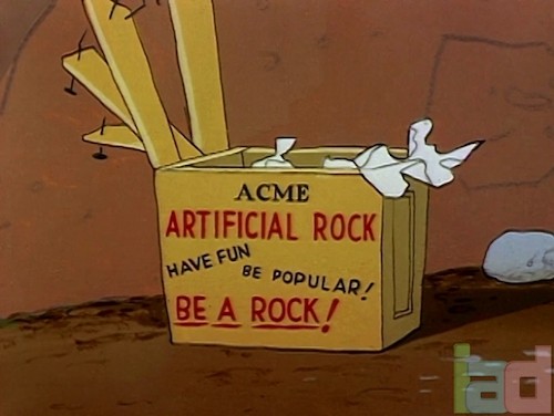 image via The Internet Animation Database, intanibase.com: screenshot from a Roadrunner cartoon showing an unpacked wooden crate labeled ACME ARTIFICIAL ROCK / HAVE FUN / BE POPULAR! / BE A ROCK!