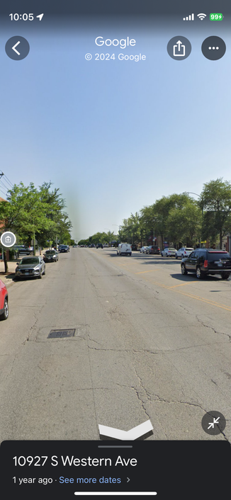 A four-lane street screenshot of Google street view. There are cars driving in the road and parked along the side. Most of the image is asphalt, and some small trees. The sky is blue.