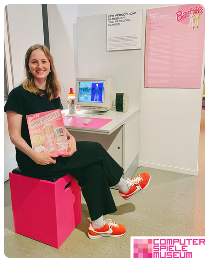 Auf einem pinken Würfelhocker sitzt eine Frau und hält die Verpackung vom PC-Spiel Barbie Mode Designer. Neben ihr steht ein Tisch mit 1990er Monitor, Tastatur, pinker Mausmatte und Lautsprechern. Auf dem PC läuft das Spiel Barbie Mode Designer. An einer Wand dahinter hängt eine große Tafer in pink und mit weißer Schrift, auf der eine Erklärung zum Spiel drauf steht.