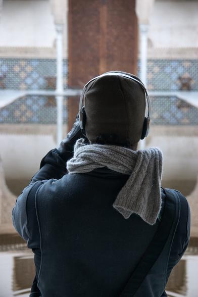 An unidentifiable person, wearing a jacket, a grey scarf, a leather hat and headphones lifts his gloved hand to take a photograph of the Alhambra, in Granada, Spain.